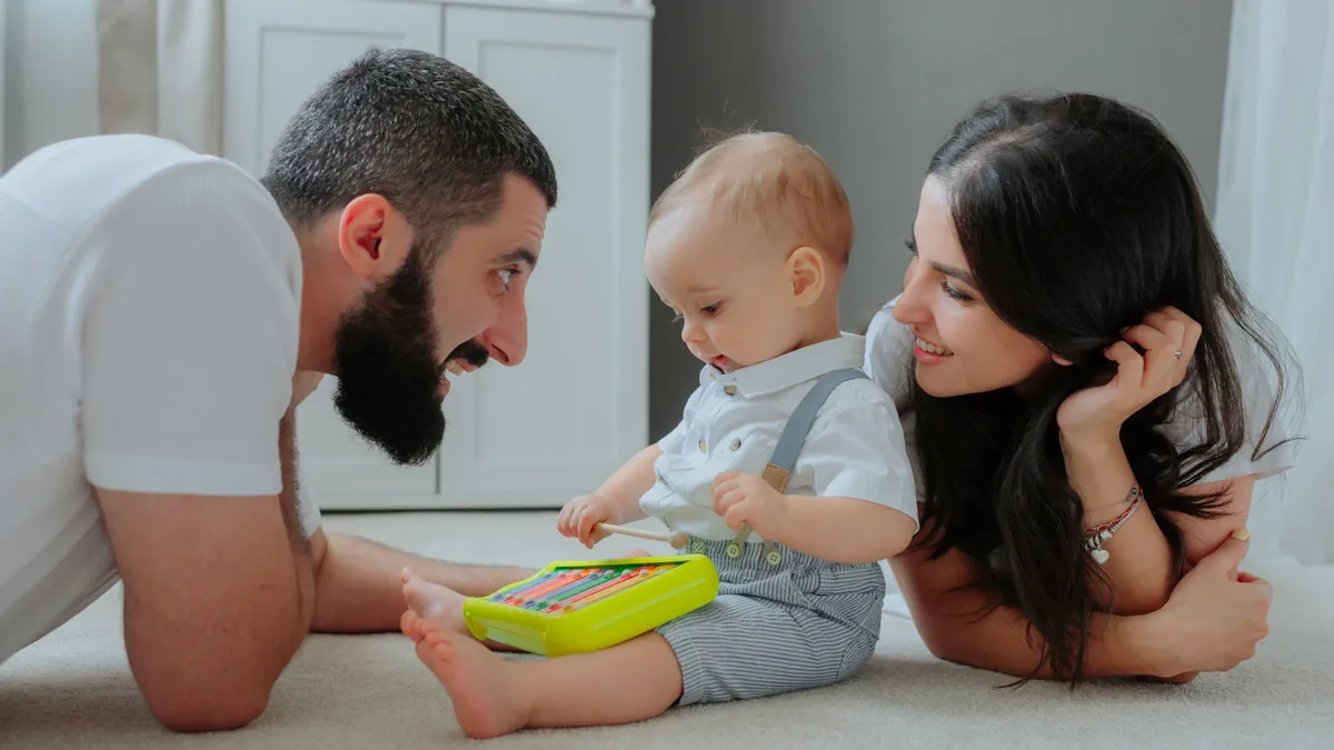 Two parents lying on the floor, smiling at their baby playing with a toy abacus.